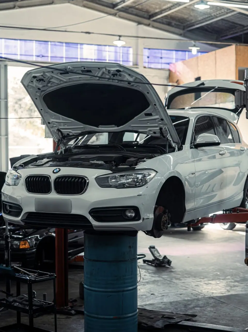 BMW Série 1 branca no elevador da oficina, com capô aberto e sem a roda dianteira